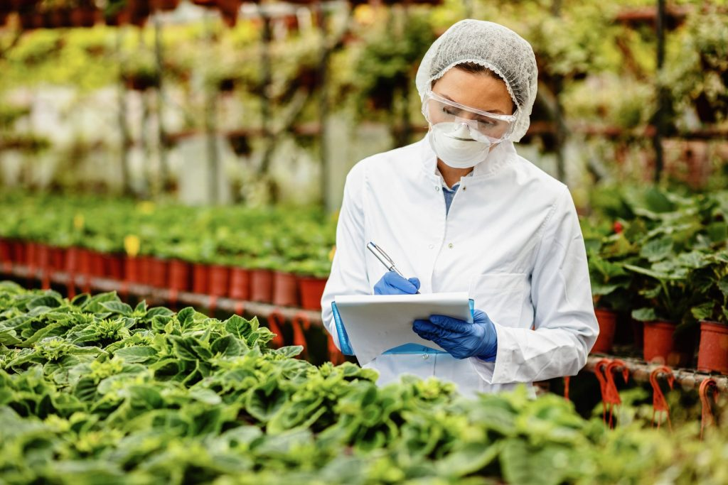 A female biotechnologist wearing a mask, gloves and a white robe is inspecting potted plants in plant nursery and writing notes into clipboard